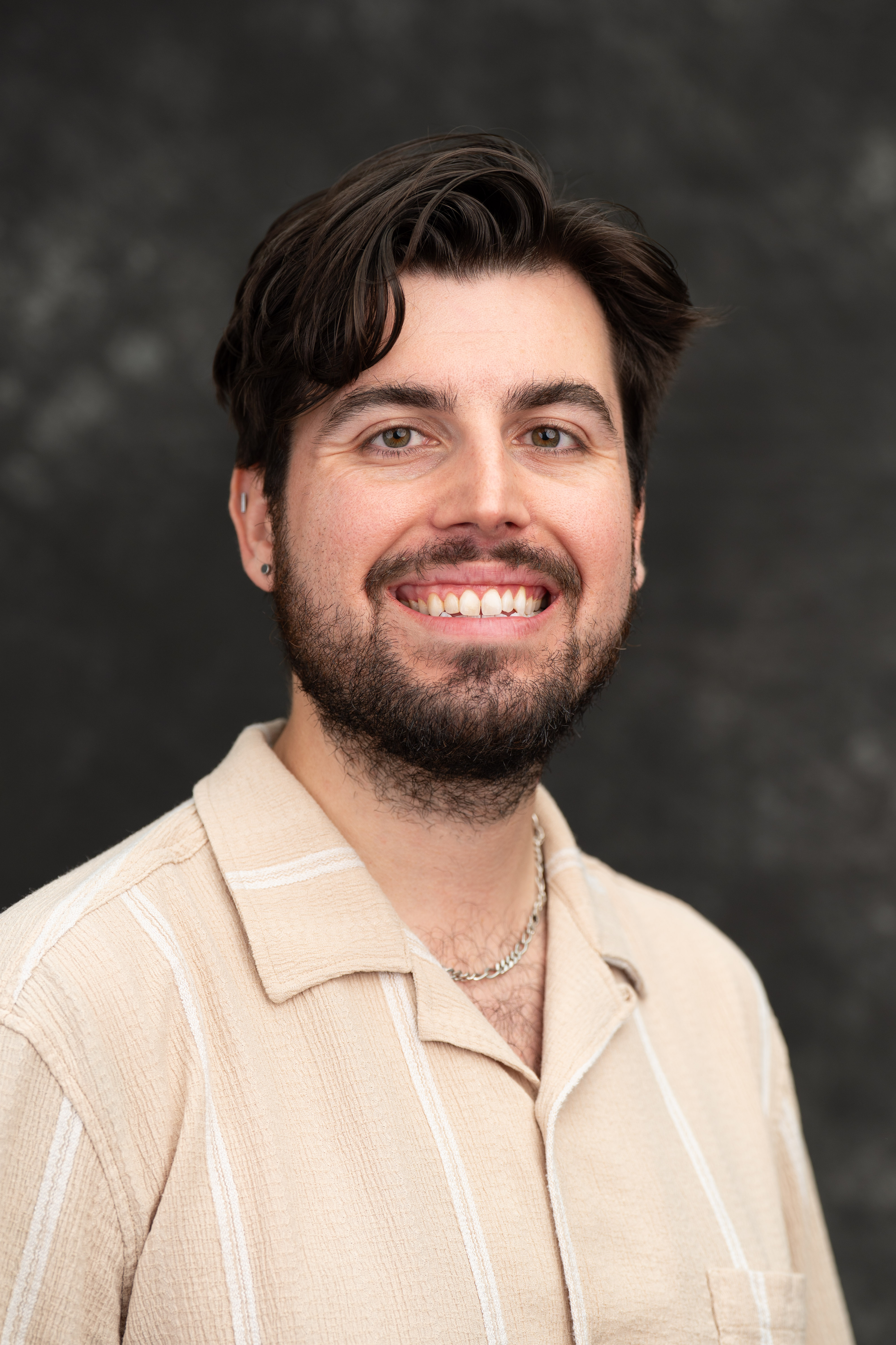 Studio headshot of Remy Beauregard, a young man with wavy dark brown hair and a dark beard, smiling at the camera, wearing a cream striped collared shirt against a mottled gray backdrop