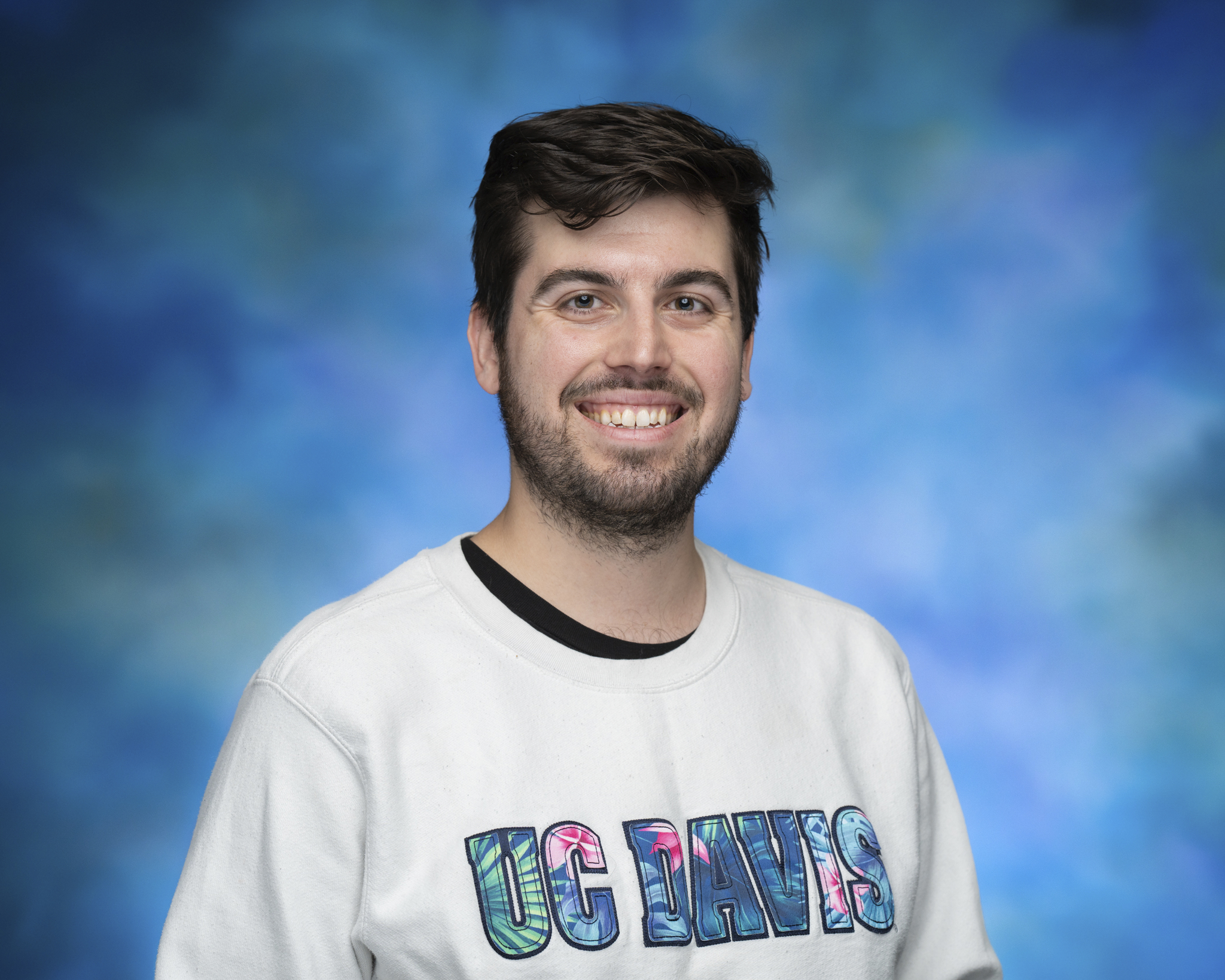 Headshot of a young man with short brown hair, smiling warmly, wearing a white UC Davis crewneck sweater against a blurred blue background