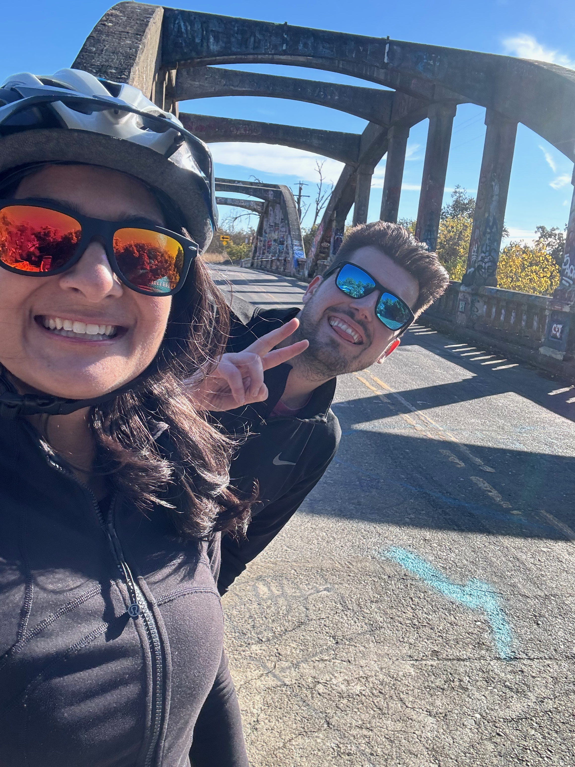 Mitali Mathur (foreground, bike helmet and sunglasses) and Remy Beauregard (background, making a peace sign) smiling on a bridge on a sunny day.